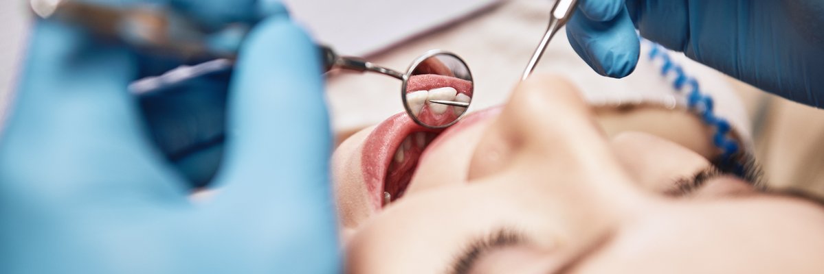 Frau mit geöffnetem Mund bei einer Zahnbehandlung. Zu sehen sind zwei Hände mit blauen Handschuhen und zahnmedizinischen Geräten bei der Untersuchung