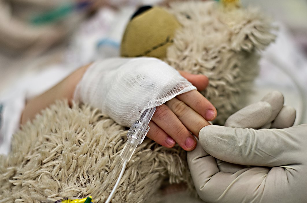 Verbundene Kinderhand am Tropf umfasst einen Teddy. Eine größere Hand im Handschuh berührt die Kinderfinger