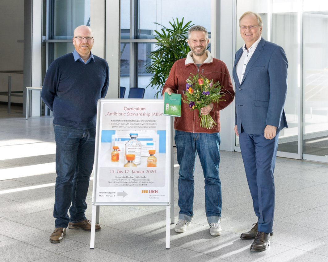 Drei Männer an einem Plakat. Einer der Männer hält einen Blumenstrauß.