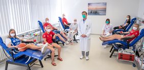 Sechs Sportlerinnen und Sportler liegen und sitzen auf Stühlen oder Liegen und spenden Blut. Die Liegen befinden sich rechts und links im Bild; in der Mitte sind Dr. Julian Hering, Leiter der Blutspende am UKH, sowie zwei Pflegekräfte zu sehen.