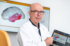 Ein weißer Mann mit Kittel und Brille deutet mit einem Kulli auf ein Tablet in seiner Hand. Darauf ist eine Präsentation abgebildet, die ein Gehirn und Datenbalken zeigt. Der Temporallappen des Gehirns ist rot eingefärbt. Die Präsentation ist auch auf einem großen Bildschirm in Hintergrund unscharf zu erkennen.