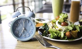 Ein blauer Metallwecker zeigt die Uhrzeit 7 Uhr. Daneben stehen ein Gemüse-Teller mit Tomaten, Broccoli, Salaten und Sprossen. Dafür liegt eine Gabel. 