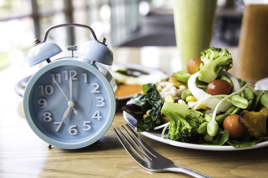 Ein blauer Metallwecker zeigt die Uhrzeit 7 Uhr. Daneben stehen ein Gemüse-Teller mit Tomaten, Broccoli, Salaten und Sprossen. Dafür liegt eine Gabel. 