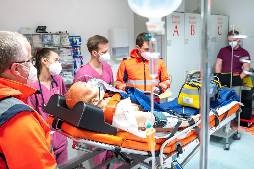 Schockraum in der Notaufnahme: Rettungssanitäter und Ärzt:innen stehen neben einer Simulationspuppe auf einer Trage. Die Puppe wird beatmet. Medizinische Geräte sind zu sehen.