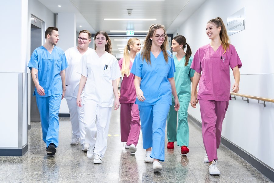 Eine Gruppe junger Menschen in medizinischer Arbeitskleidung in verschiedenen Farben läuft miteinander redend einen Gang entlang.  Eine Gruppe junger Menschen in medizinischer Arbeitskleidung in verschiedenen Farben läuft miteinander redend einen Gang entlang.