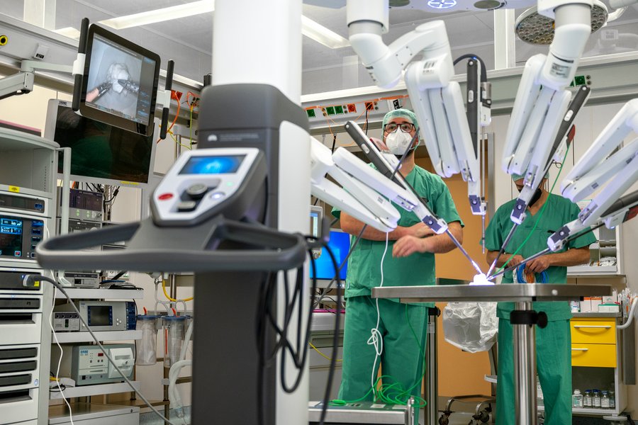 Zwei Männer in grüner OP-Kleidung stehen an den Roboterarmen eines OP-Robotersystems. Im Hintergrund ist ein Bildschirm zu sehen. Das Foto zeigt einen OP-Saal  Zwei Männer in grüner OP-Kleidung stehen an den Roboterarmen eines OP-Robotersystems. Im Hintergrund ist ein Bildschirm zu sehen. Das Foto zeigt einen OP-Saal