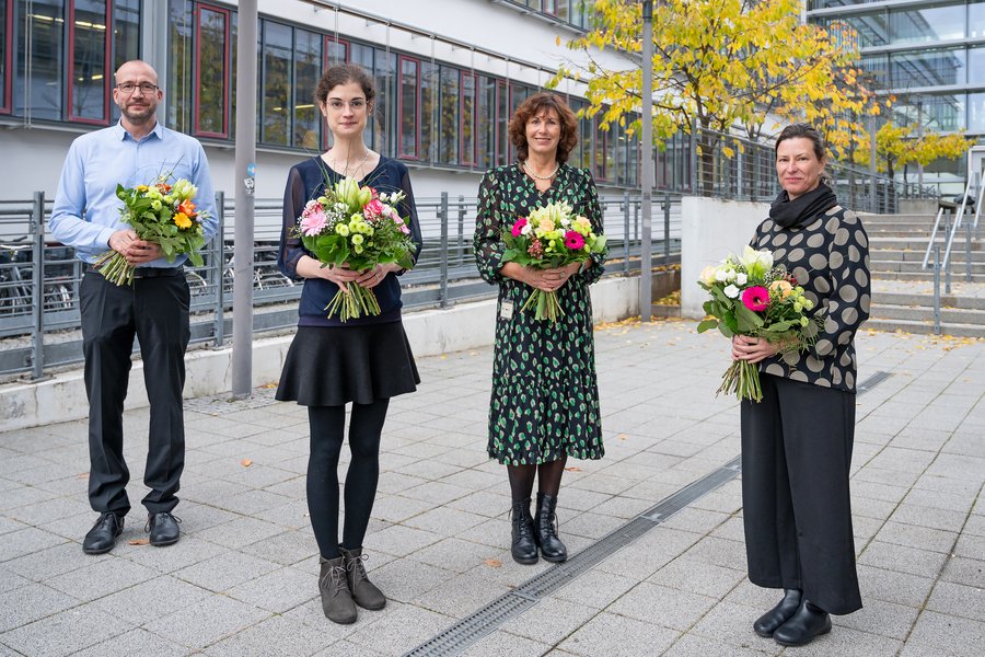 Ein Mann und drei Frauen stehen vor dem Lehrgebäude am Universitätsklinikum. Alle vier haben bunte Blumensträuße in der Hand. 