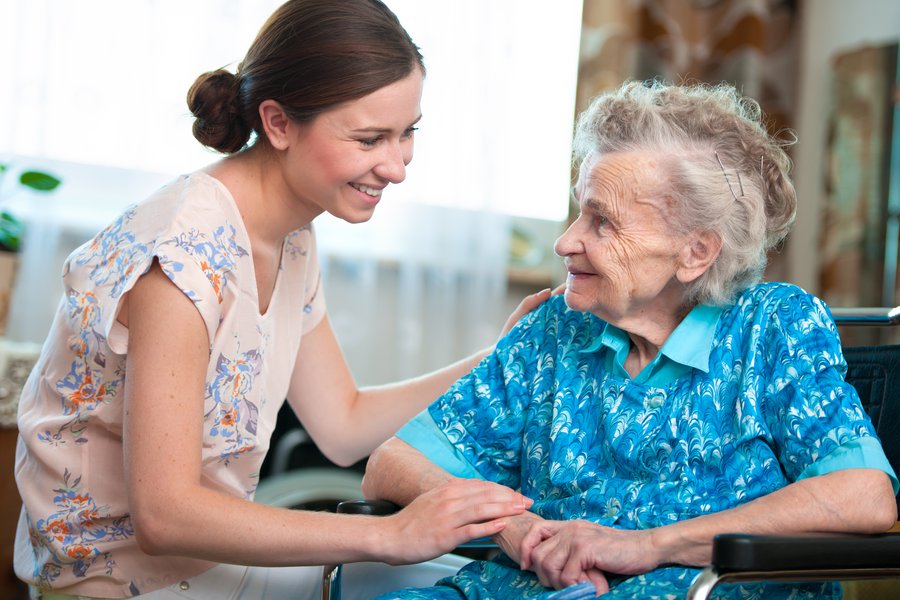 Eine junge Frau links im Bild, braune Haare hält die Hand einer Seniorin, die in einem Rollstuhl sitzt.
