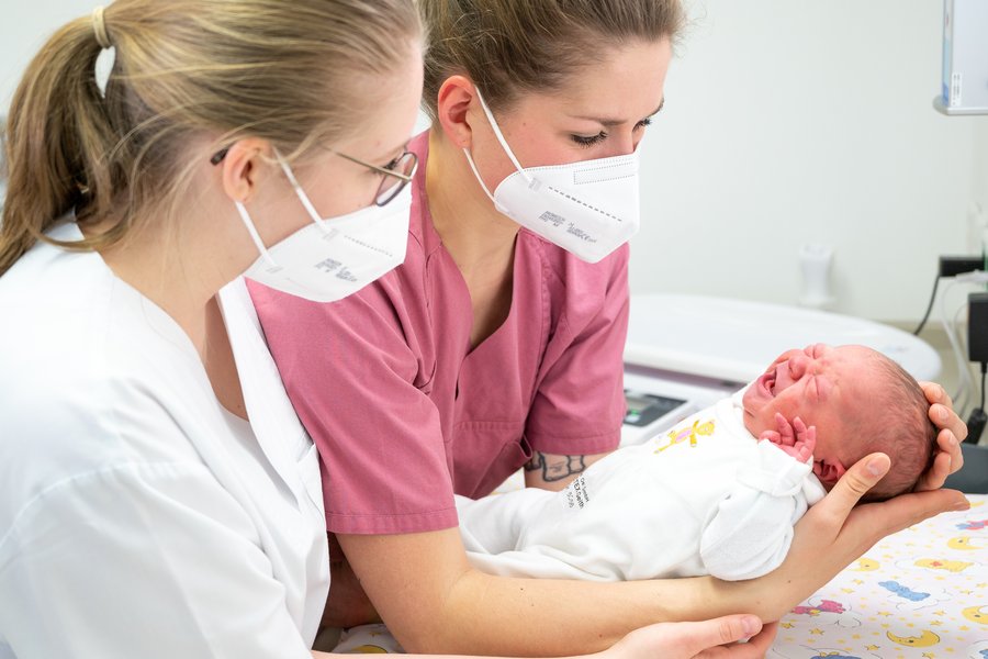 Zu sehen sind zwei Frauen in Klinikkleidung, die ein Neugeborenes halten.  Zu sehen sind zwei Frauen in Klinikkleidung, die ein Neugeborenes halten.
