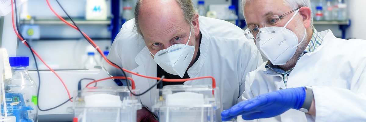 Zwei Männer mit Masken und weißen Kitteln schauen über eine Laborstrecke mit einer Experimentieranordnung