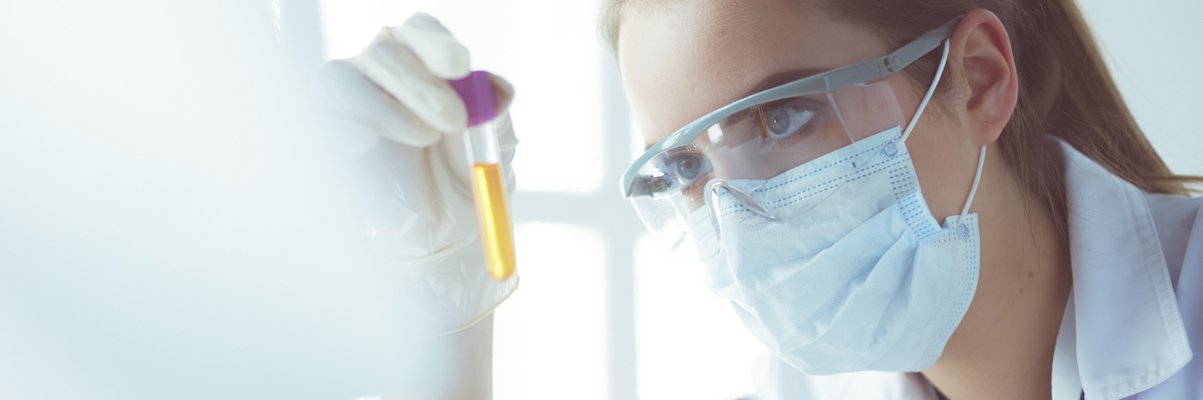 Frau im weißen Kittel mit Schutzbrille und Maske schaut auf eine Reagenzglas mit einer gelben Flüssigkeit. Vor ihr stehen weitere Proben.