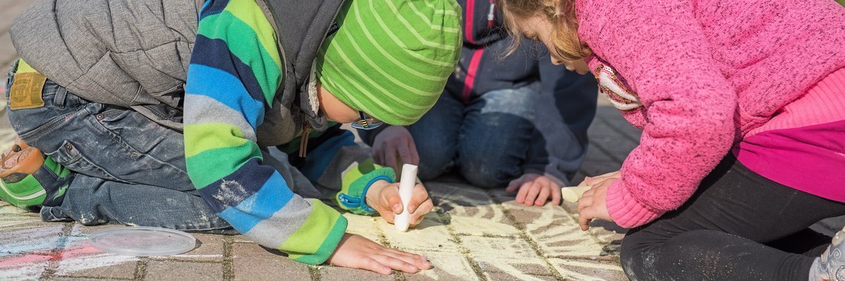 Drei Kinder malen mit Kreide auf den gepflasterten Weg.