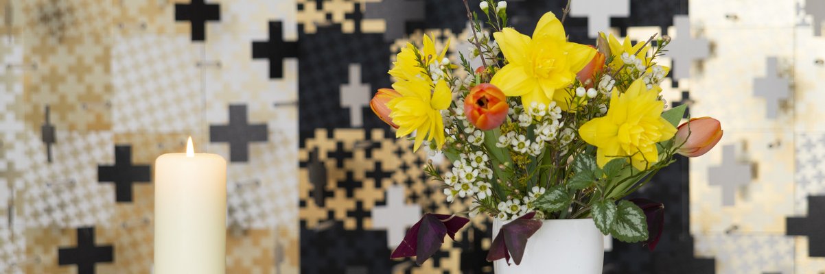 Eine brennende weiße Kerze und ein Strauß Frühlingsblumen stehen auf einem Glastisch. Im Hintergrund ist eine gemusterte Tapete in Schwarz, Weiß und Gold zu sehen.