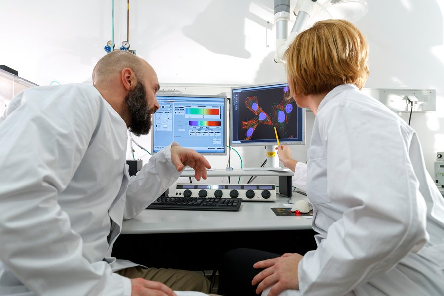 Ein Mann mit Bart und Glatze und eine Frau mit blondem Haar blicken im Labor auf mehrere Bildschirme mit Daten.