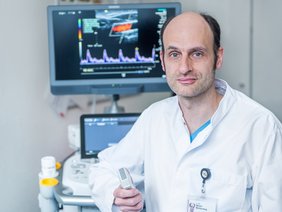 Auf dem Foto ist ein Mann in weißen Arztkittel. Er hält ein Ultraschallgerät in der rechten Hand, im Hintergrund sind Monitore zu sehen.