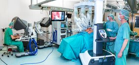 Blick in einen OP-Saal mit dem da Vinci-Roboter, dessen Arme über einem Patienten schweben. Man sieht Chirurgen, die an Konsolen den Roboter bedienen, ein Chirurg steht neben dem Patienten. Vom Patienten ist nur der Bauch zu sehen, der für die Operation aufgebläht ist. Auf einem Monitor ist das Innere des Bauchraums zu sehen.