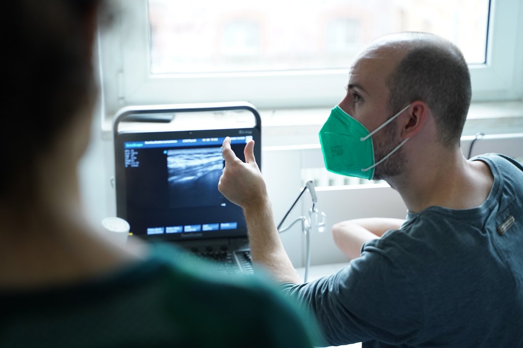 Ein Mann schaut links zur Seite, mit Daumen und Zeigefinger deutet er etwas an, das er zeigt. Vor ihm steht ein kleiner Monitor, auf dem ein Ultraschall-Bild zu sehen ist.