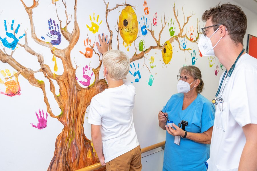 Ein junge mit blonden Haaren und weißem T-Shirt interlässt einen blauen Handabdruck an einem gemalten Baum an der Wand. Neben ihm steht ein Arzt und eine Pflegefachfrau.