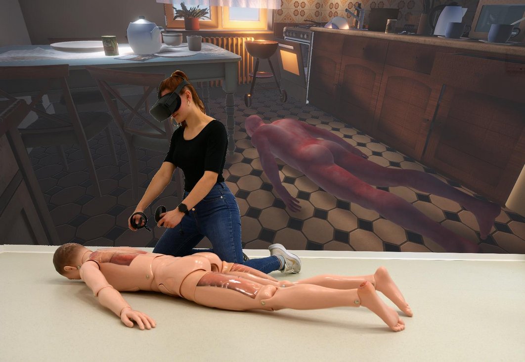 Virtual Reality: Eine Frau mit VR-Brille kniet neben einer Simulations-Puppe, die bäuchlings auf dem Boden liegt. Im Hintergrund ist ein 3D-Animation zu sehen, in der eine Person bäuchlings in einer Küche liegt.