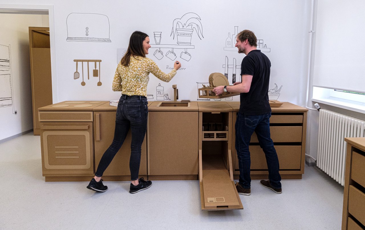 Zwei Personen simulieren eine Situation in einer Küche aus Pappelementen.