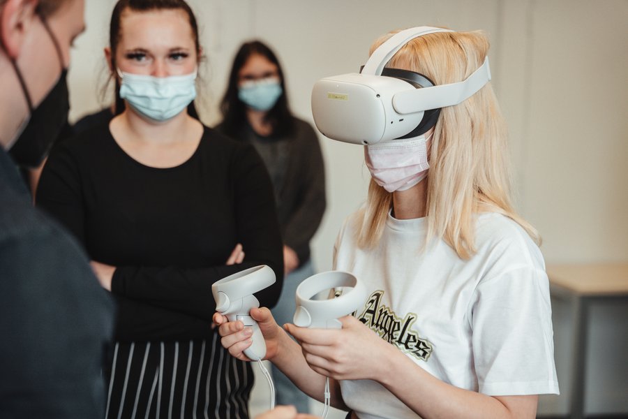 Im rechten Bildteil ist eine junge Frau zusehen, die eine VR-Brille trägt und zwei Controller in ihren Händen hält. Im linken Bildteil stehen drei weitere Personen mit Mundschutz und beobachen sie dabei. Sie sind nicht scharf zu erkennen.