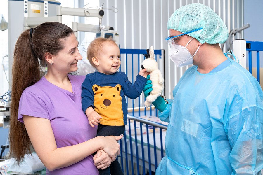 Eine junge Frau hat ein kleines Kind auf dem Arm. Ein Arzt in medizinscher Schutzkleidung steht rechts von ihnen und reicht dem Kind einen teddybär.