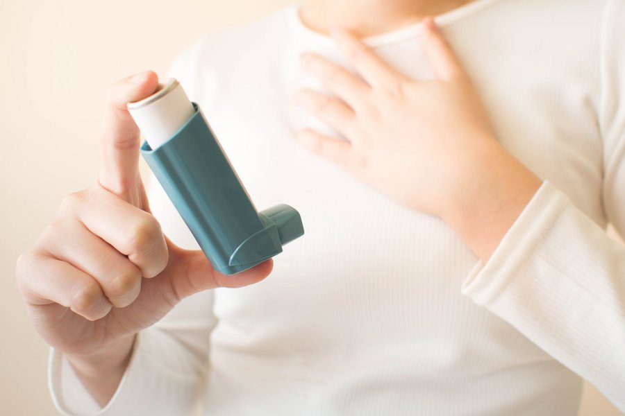 Ein Kind hält einen Asthma-Inhalator in der einen Hand, mit der anderen hält es sich die Brust. Nur der Oberkörper ist zu sehen. 