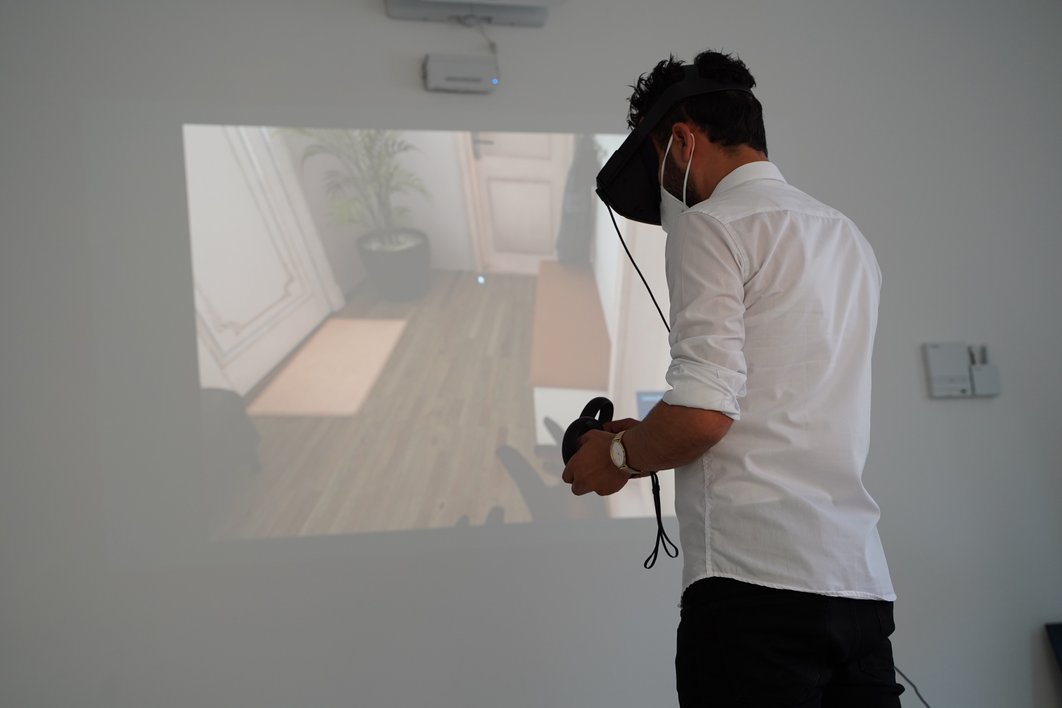 Ein Mann mit VR-Brille ist von schräg hinten zu sehen. An der Wand vor ihm ist ein projiziertes Bild zu sehen, das einen Wohnbereich zeigt.