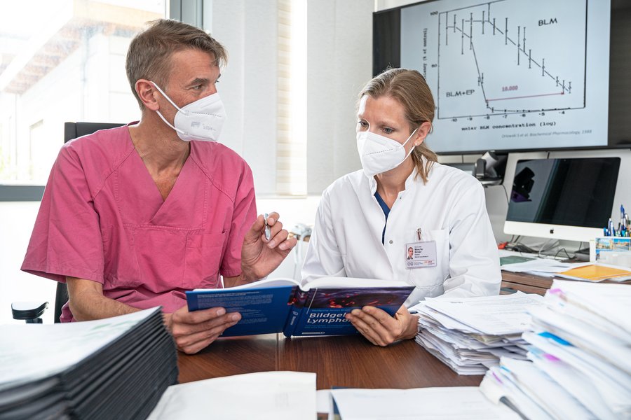Ein Mann links mit brombeerfarbener medizinischer Arbeitskleidung und eine Frau rechts in weißem Arztkittel schauen gemeinsam auf ein Tablet. Sie sitzen an einem Schreibtisch