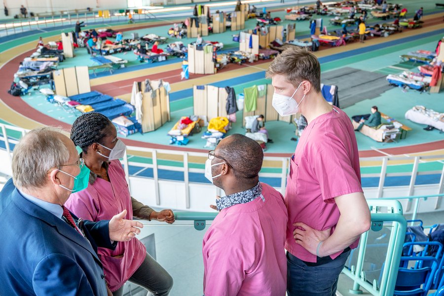Drei Männer und eine Frau in medizinscher Arbeitskleidung (brombeerfarben) sind im Gespräch zu sehen. Im Hintergrund ist eine Sporthalle mit Feldbetten zu sehen.