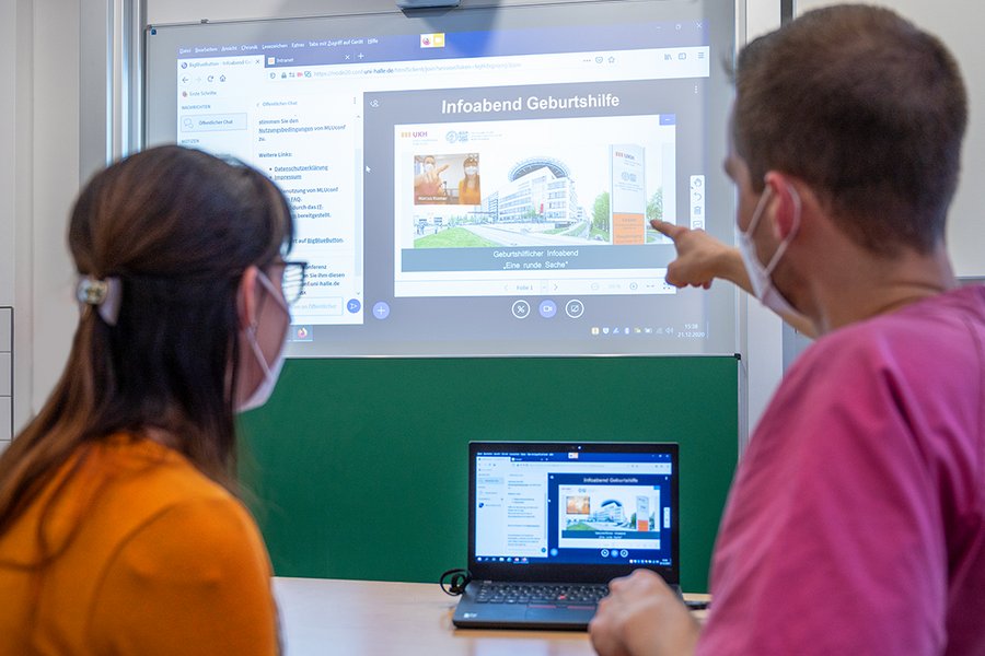 Eine Mitarbeiterin und ein Mitarbeiter sitzen vor einem Laptop. Sie sind von hinten zu sehen. Der Mitarbeiter zeigt auf eine Leinwand im Hintergrund, auf der eine Folie mit der Aufschrift "Infoabend Geburtshilfe" zu sehen ist.