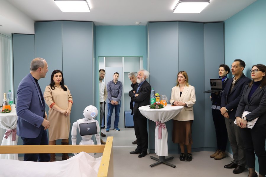 Ein modern wirkender Raum mit zehn Menschen. In der Mitte steht ein Kommunikationsroboter mit dem Namen "Pepper".  Ein modern wirkender Raum mit zehn Menschen. In der Mitte steht ein Kommunikationsroboter mit dem Namen "Pepper".