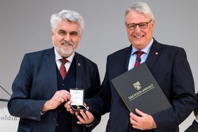 Zwei Männer mit grauem Haar und in dunklen Anzügen blicken in die Kamera. Links hält eine Ehrennadel, rechts eine Mappe