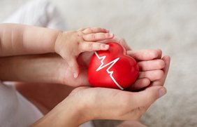 Nahaufnahme von Händen von Baby und Eltern, die ein rotes Herz festhalten, auf dem eine EKG-Linie abgebildet ist.