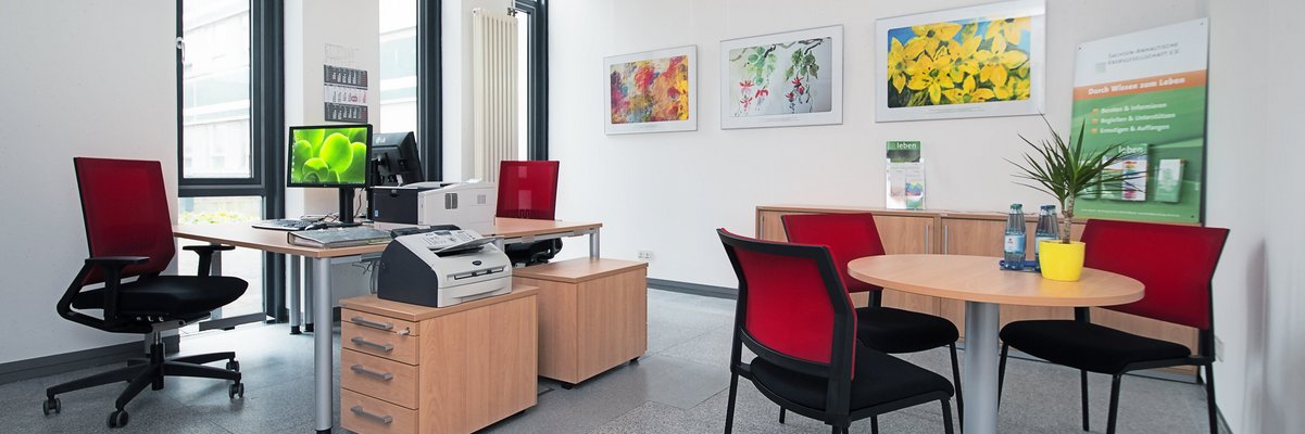 Blick in ein Beratungszimmer mit Tischen, Computern und roten Stühlen