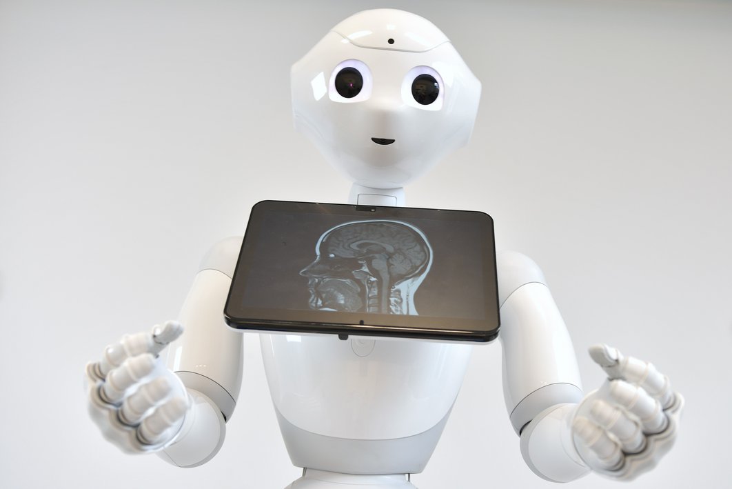 Weißer menschenähnlicher Roboter schaut mit großen Augen in die Kamera und zeigt auf einen Bildschirm unterhalb seines Kopfes. Darauf zu sehen ist die Röntgen-Aufnahme eines menschlichen Schädels im Profil.
