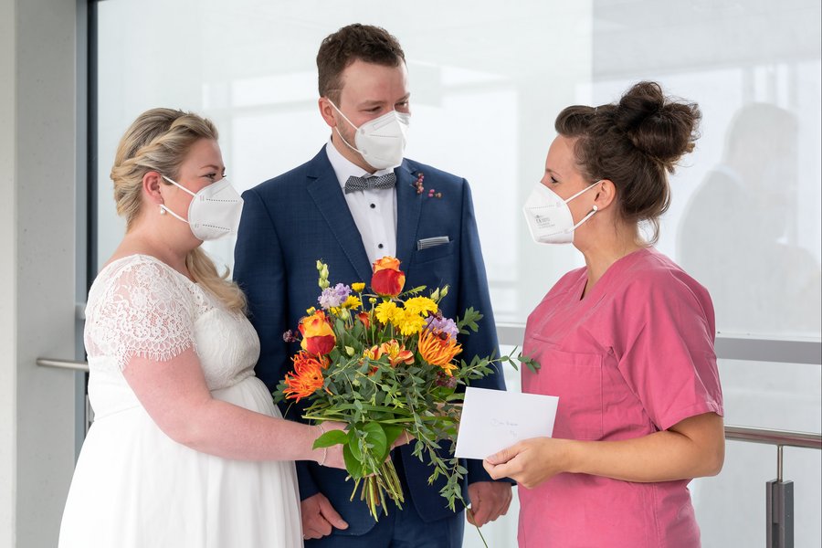 zu sehen ist links eine Braut in Brautkleid, in der Mitte ein Bräutigam im Anzug. Rechts überreicht eine Frau in medizinischer Arbeitskleidung (magenta) einen Blumenstrauß und einen Umschlag.