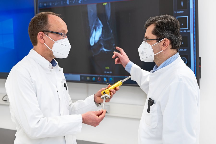 zwei Männer in weißen Arztkittel stehen vor einem Monitor, auf dem ein Röntgenbild zu sehen ist. Der Mann auf der linken Seite hält ein künstliches Kniegelenk in der Hand