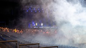Ein großer Konzertsaal mit schummrigem Licht, einer Bühne, auf der Menschen stehen, von rechts kommt weißer Nebel ins Bild.