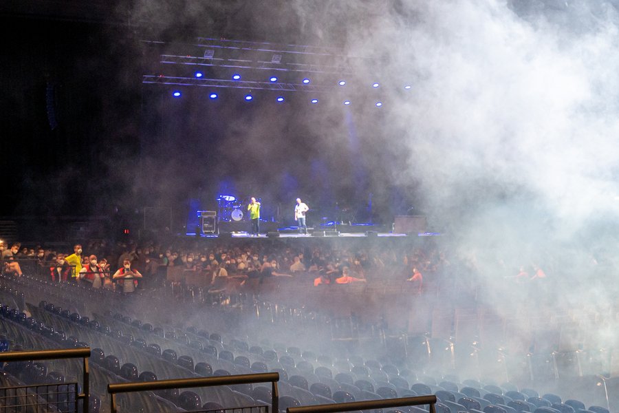 Ein großer Konzertsaal mit schummrigem Licht, einer Bühne, auf der Menschen stehen, von rechts kommt weißer Nebel ins Bild.