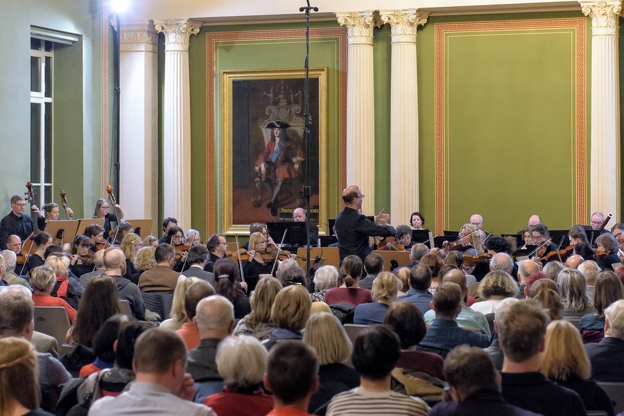Orchester bei einem Konzert aus Zuschauerperspektive, in der Mitte stehend der Dirigent mit dem Rücken zum Publikum