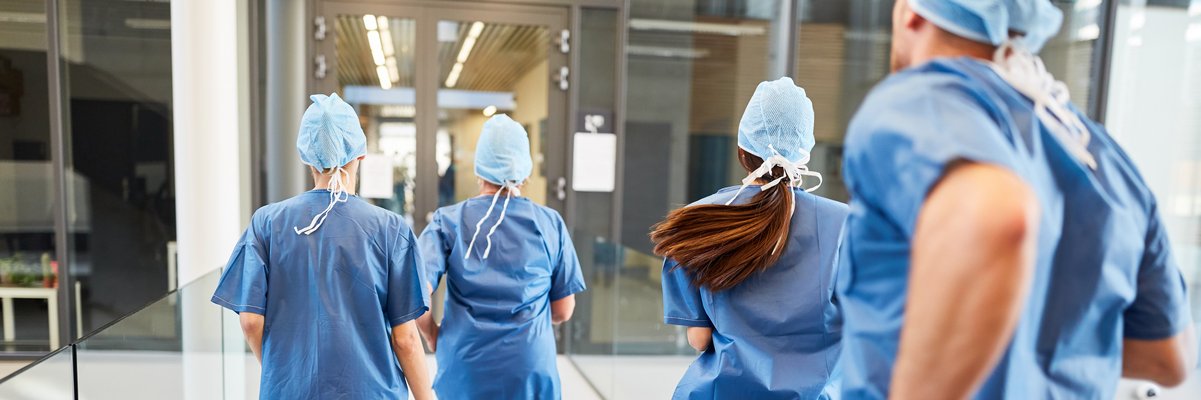 Medizinisches Personal in blauer Arbeitskleidung mit Maske und Haube rennt über den Klinikgang.